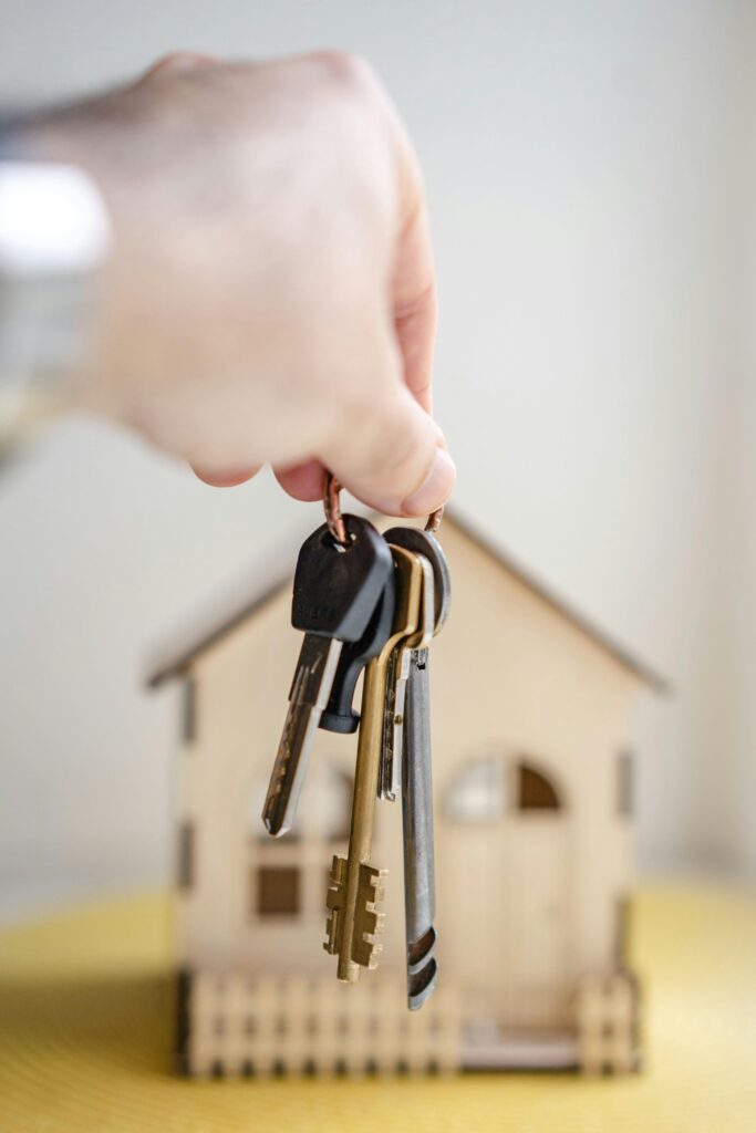 Close-up of a hand holding keys with a miniature wooden house in the background, symbolizing real estate investment.